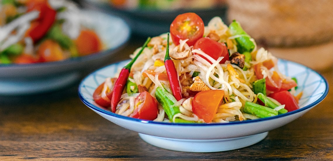 An overhead view of a colorful plate of Som Tam, surrounded by ingredients including green papaya, cherry tomatoes, green beans, peanuts, lime wedges, and chili peppers, on a wooden table.