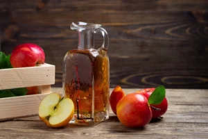 Botol cuka sari apel transparan dengan label organik diletakkan di atas meja kayu, dengan apel segar di sampingnya sebagai dekorasi